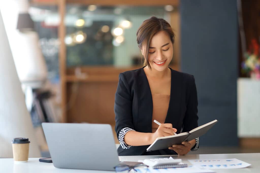 Beautiful Asian businesswoman using her laptop to work and enjoy taking notes on meeting minutes agendas of secretarial work.