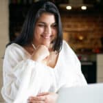 Cheerful young overweight woman listening to music while studying online using earhpones and laptop. Pretty chubby girl watching webinar or having video call on headset. Distant education concept