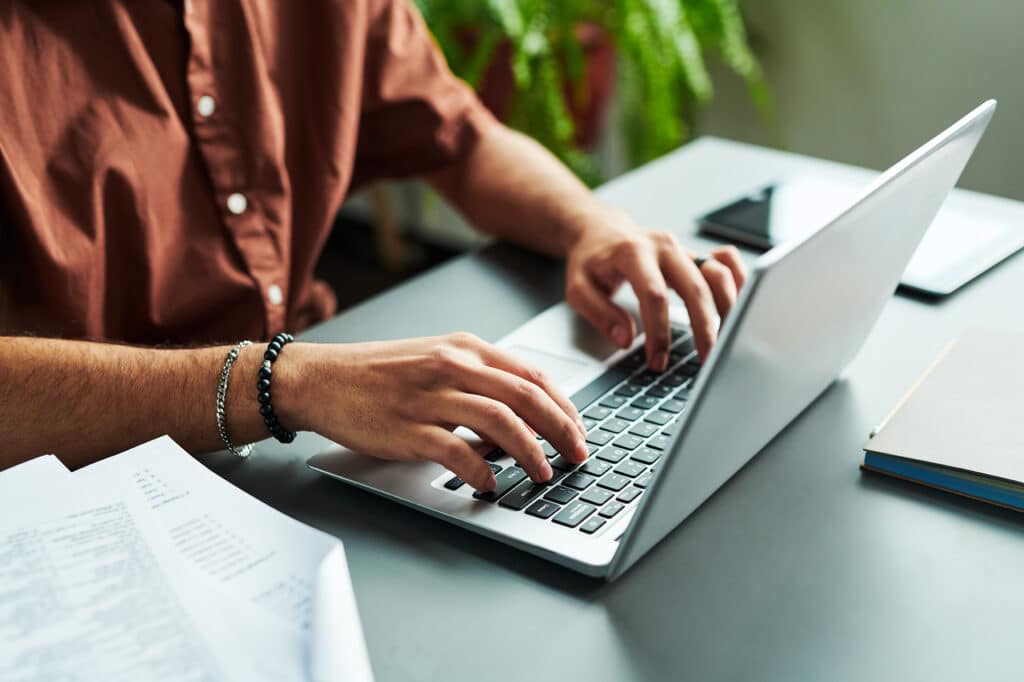 Close-up of hands of young student typing on laptop keyboard while sitting by workplace and carrying out online grammar test