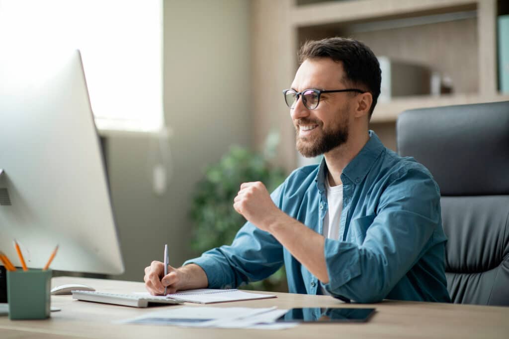 Online education, e-learning. Happy american man in stylish casual clothes, studying remotely, using a laptop, listening to online lecture, taking notes while sitting at home, smiles